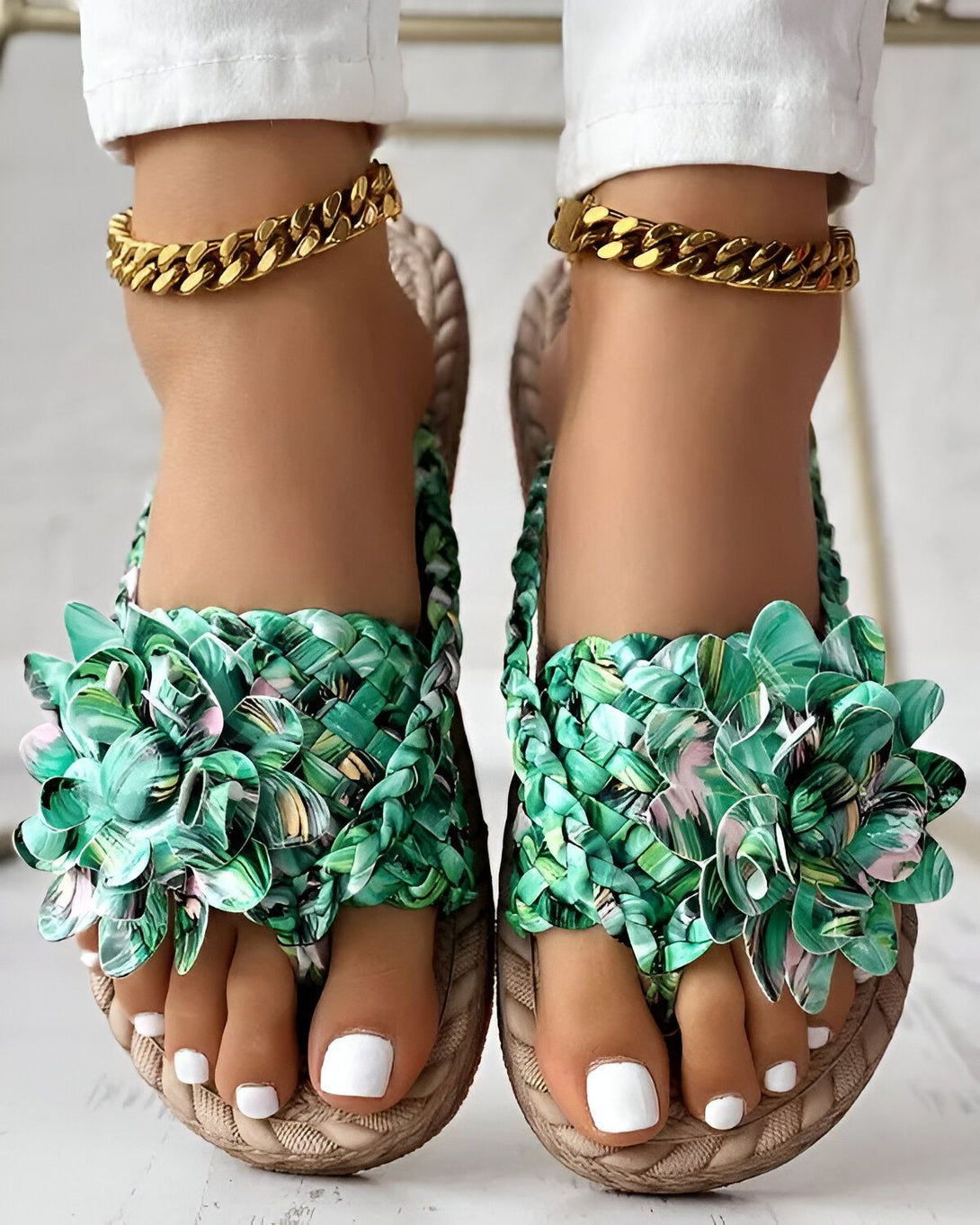 Une paire de tongs plates à enfiler, avec une bride large et des fleurs en tissu vert et rose sur le dessus. Les tongs sont portées par une femme qui a les ongles des pieds peints en blanc et qui porte un pantalon blanc et des bijoux de cheville dorés.