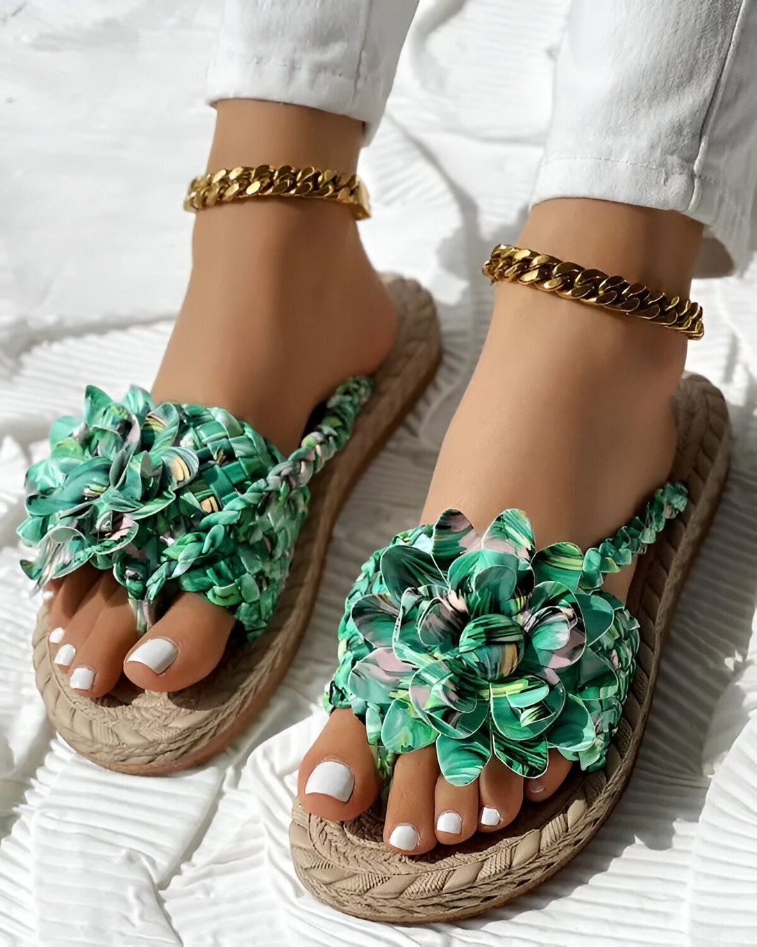 Une femme porte des sandales plates vertes à fleurs. Les sandales sont faites de tissu et ont une semelle en corde. La femme a les ongles des pieds peints en blanc et porte un bracelet de cheville doré.