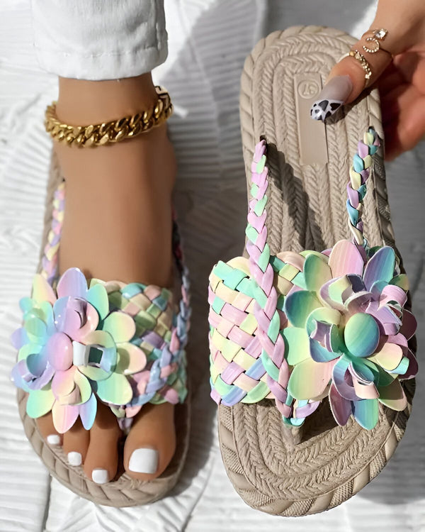 Une femme porte des sandales plates beiges avec des fleurs colorées. Les fleurs sont roses, bleues, vertes et jaunes. Les sandales sont faites de lanières tressées. La femme a les ongles peints en rose et porte un bracelet de cheville doré.