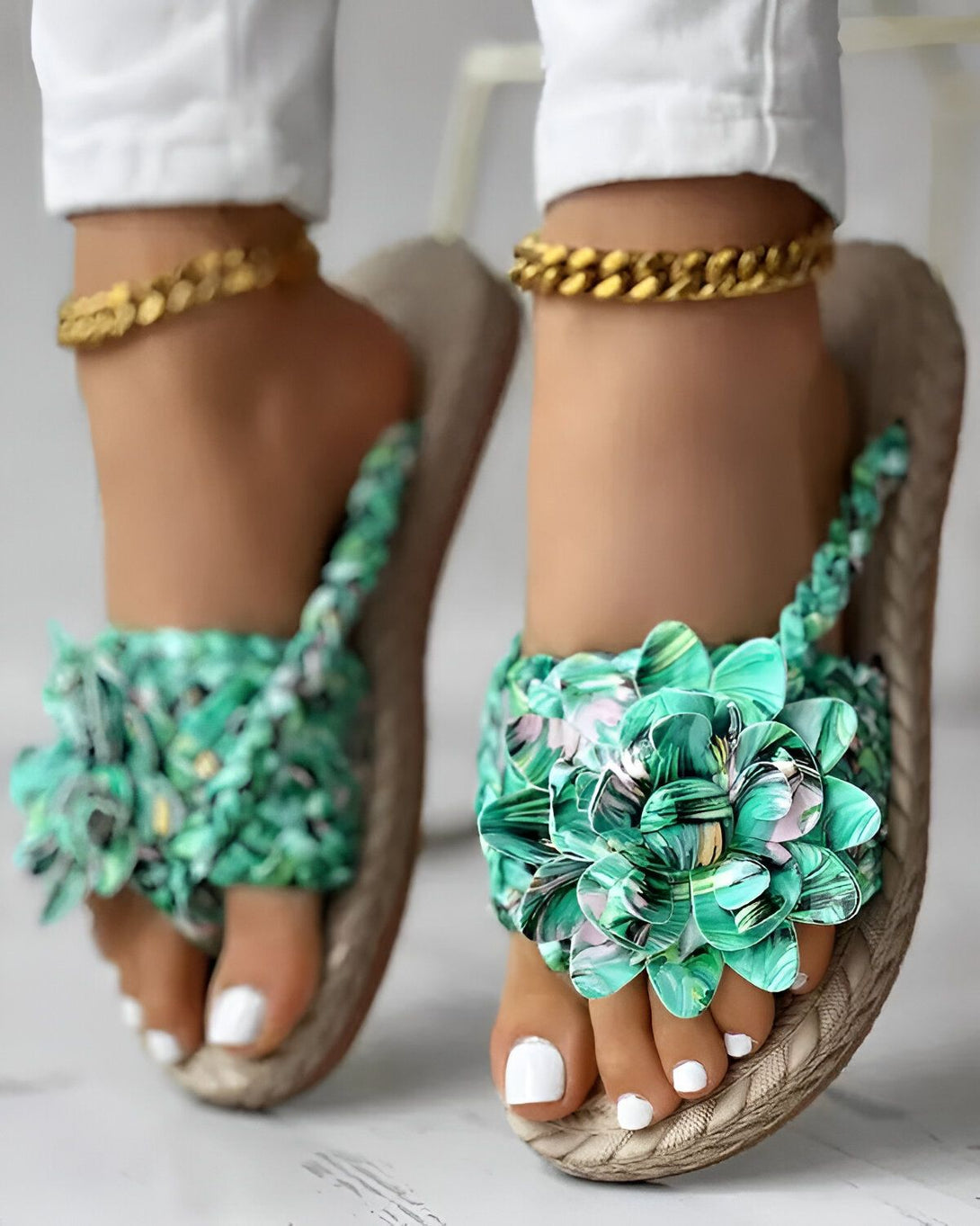 Une femme porte des sandales plates vertes à fleurs. Les sandales sont faites de tissu et ont une semelle en caoutchouc. La femme a les ongles des pieds peints en blanc et porte des bracelets de cheville en or. Elle porte également un jean blanc et un haut blanc.