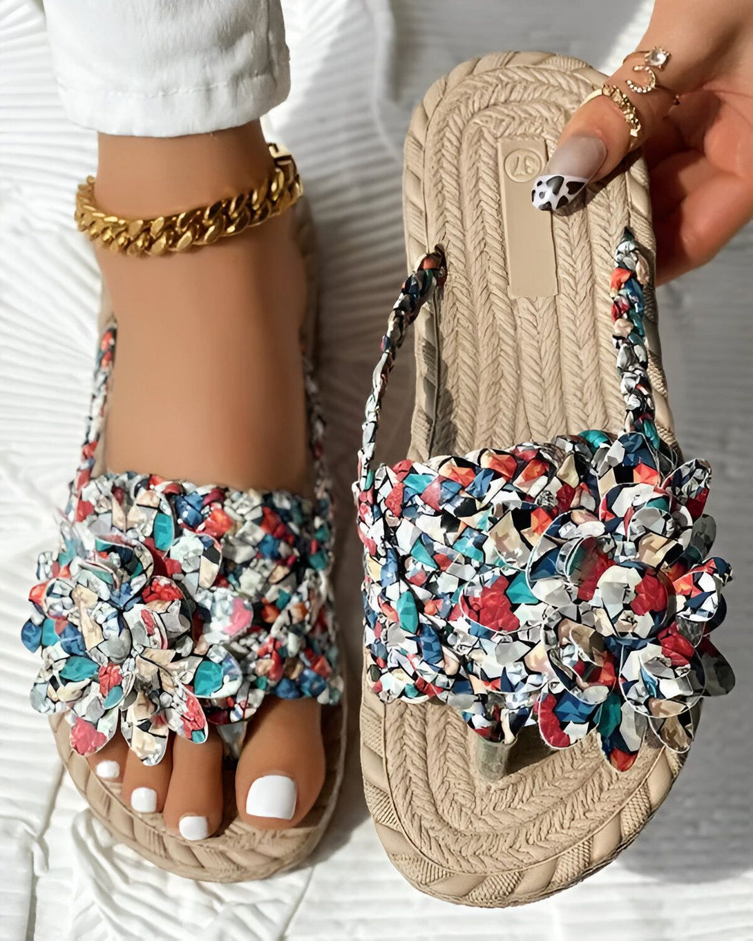 Une femme porte des sandales plates beiges à fleurs. Les sandales sont ornées de fleurs en plastique de couleur vive. La femme a les ongles des pieds peints en blanc. Elle porte un pantalon blanc et un haut blanc. Elle est debout sur un tapis blanc.