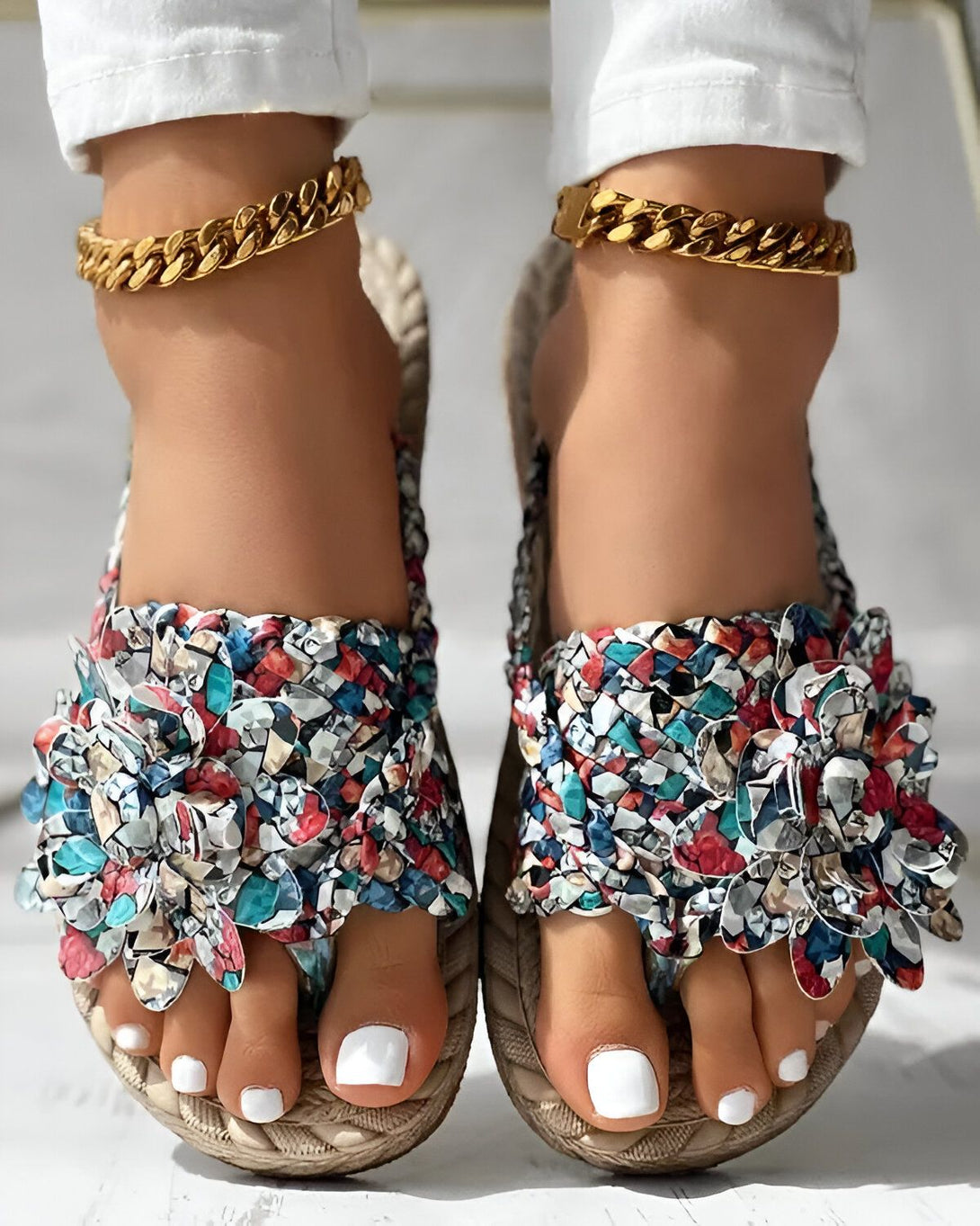 Une femme porte des sandales plates à enfiler. Les sandales sont de couleur beige et ont un motif floral coloré. La femme porte également un pantalon blanc et un haut blanc. Elle est debout sur un sol en béton.