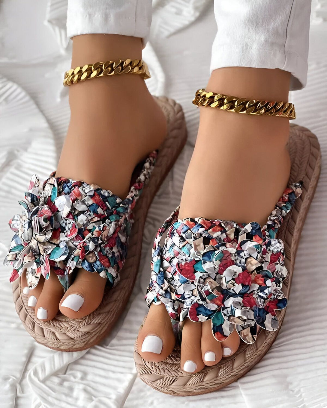 Une femme porte des sandales plates à enfiler. Les sandales sont de couleur beige et ont un motif floral coloré. La femme porte également un pantalon blanc et un chemisier blanc. Elle est debout sur un tapis blanc.