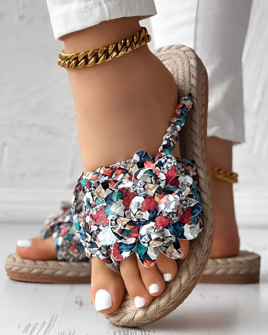 Une femme porte des sandales plates à fleurs colorées. Les sandales sont faites de paille et ont une semelle en caoutchouc. La femme porte également un pantalon blanc et un chemisier blanc. Elle a les cheveux longs et blonds et porte un collier en or. Elle est debout sur un sol en béton.