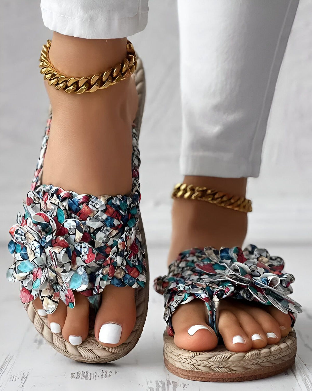 Une femme porte des sandales plates à enfiler. Les sandales sont de couleur beige et ont un motif floral coloré. La femme a les ongles des pieds peints en blanc et porte des chaînes dorées aux chevilles.