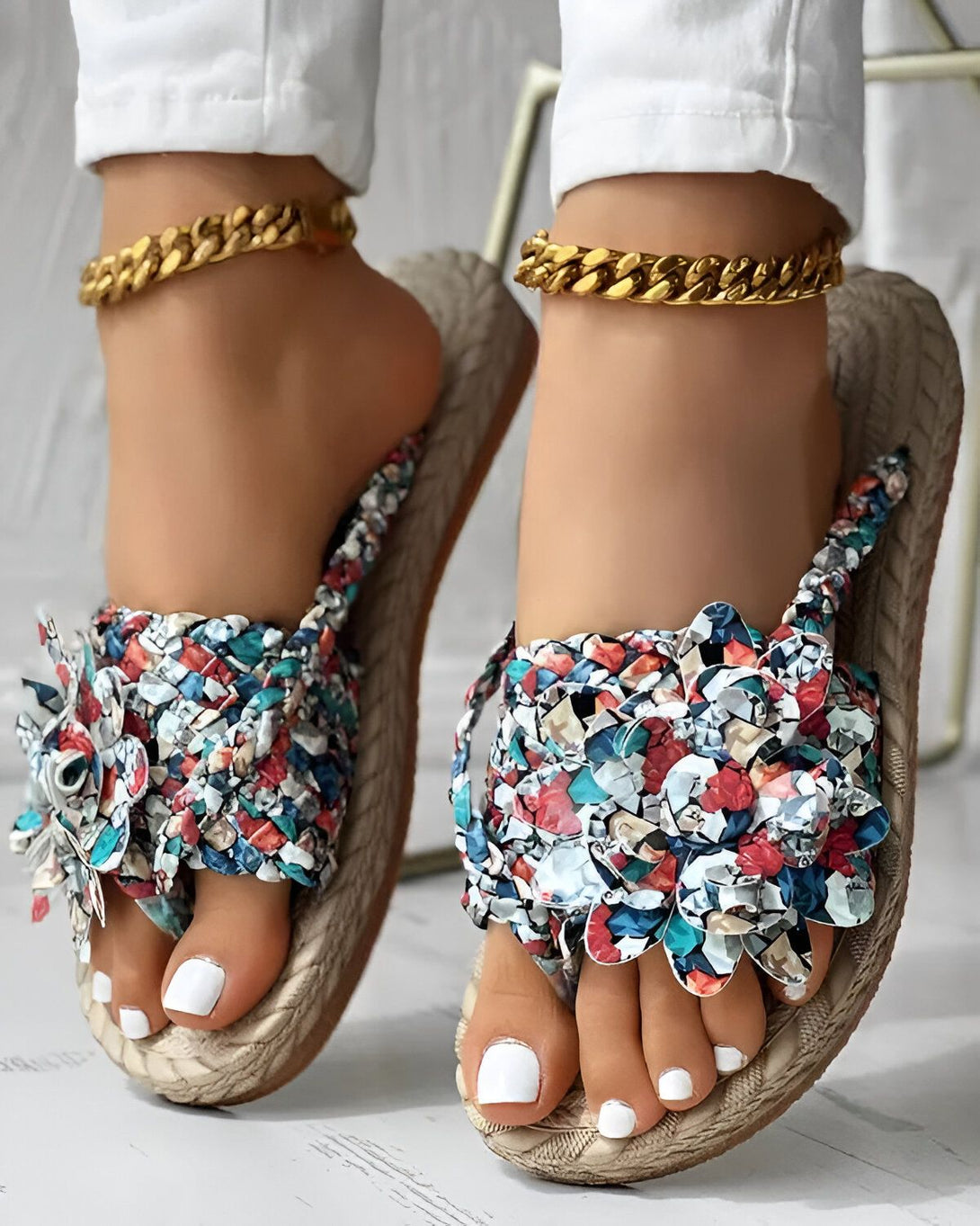 Une femme porte des sandales plates à fleurs. Les sandales sont de couleur beige et ont une bride qui passe entre les orteils. La femme porte également un pantalon blanc et un chemisier blanc. Elle est assise sur une chaise et l'on peut voir ses pieds.