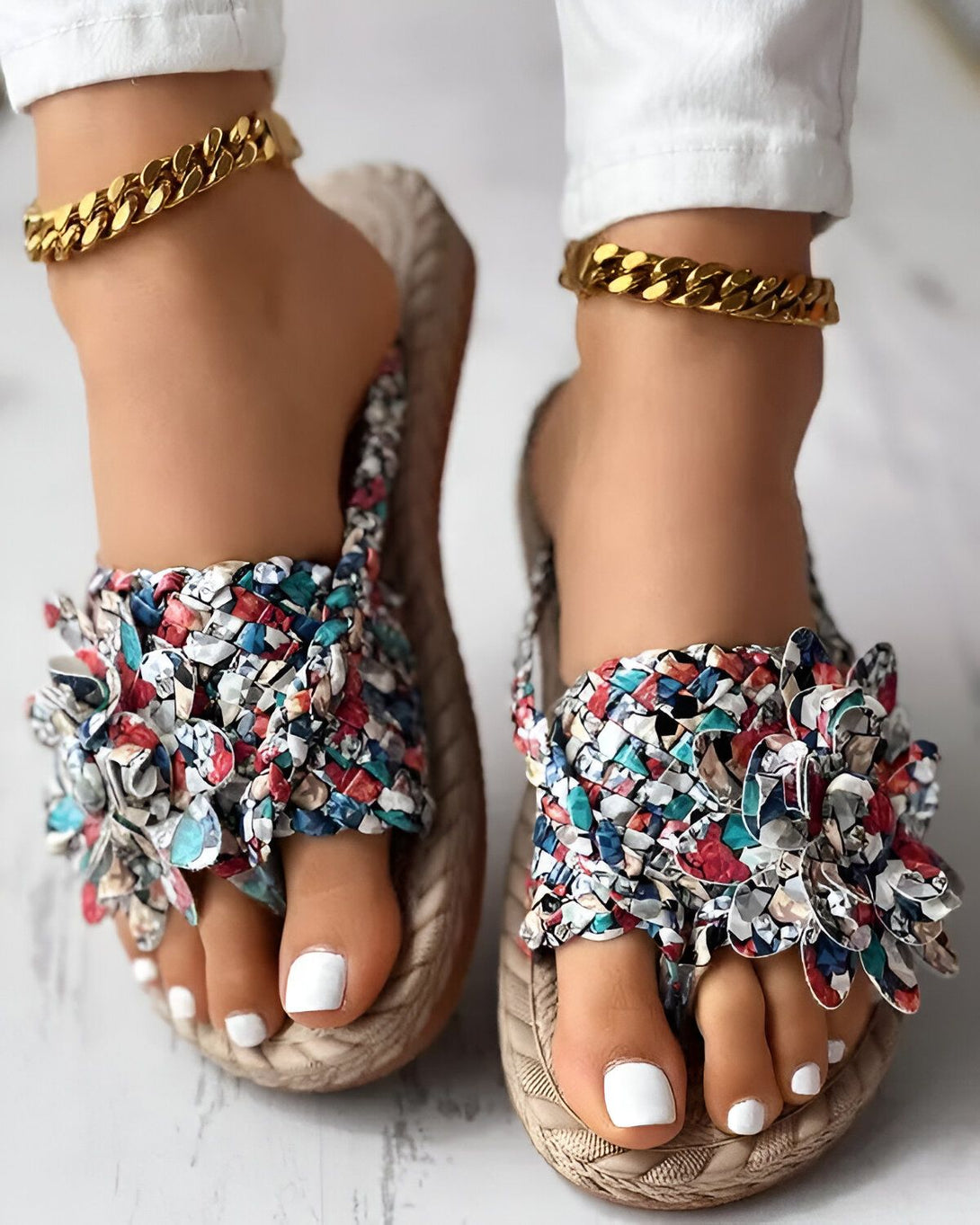 Une femme porte des tongs plates à fleurs multicolores. Les tongs sont en matières synthétique et ont une semelle compensée en corde. La femme porte également des bijoux de cheville en or.