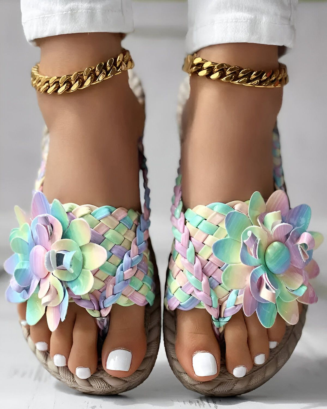 Une paire de sandales plates pour femmes avec des fleurs colorées sur le dessus. Les sandales sont de couleur beige et ont une semelle en corde. Les fleurs sont de couleur rose, jaune, bleu et vert. Les sandales sont portées par une femme avec un pantalon blanc et un haut blanc. La femme est debout sur un sol en béton.