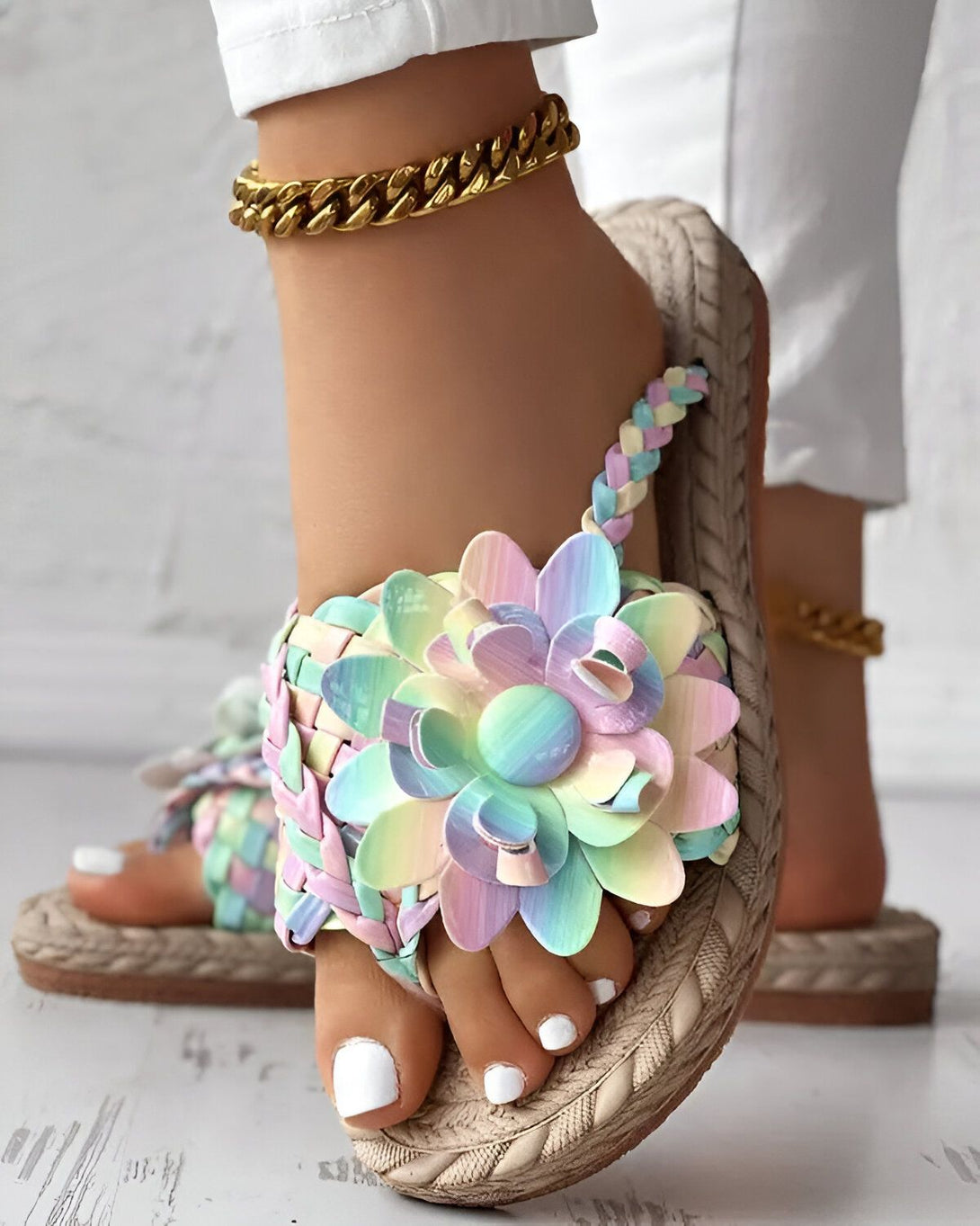 Une femme porte des sandales plates à fleurs colorées. Les sandales sont faites de cuir et ont une semelle en corde. La femme porte également un pantalon blanc et un bracelet de cheville doré.