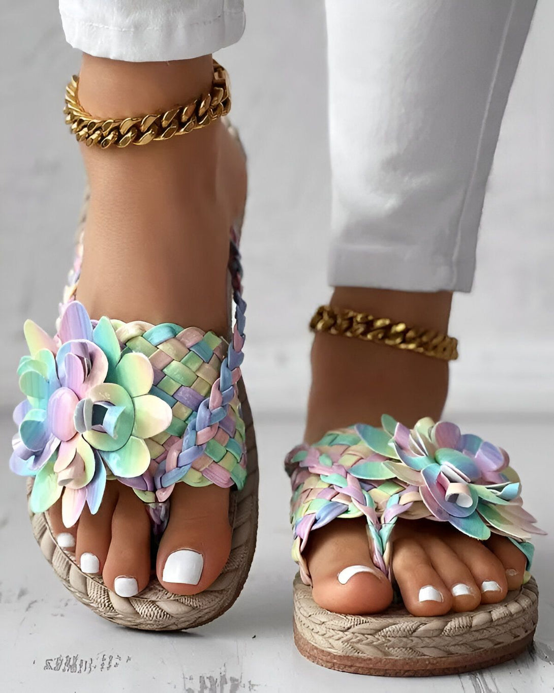 Une femme porte des sandales plates à enfiler avec des fleurs colorées sur le dessus. Les sandales sont portées avec un jean blanc et une cheville dorée.