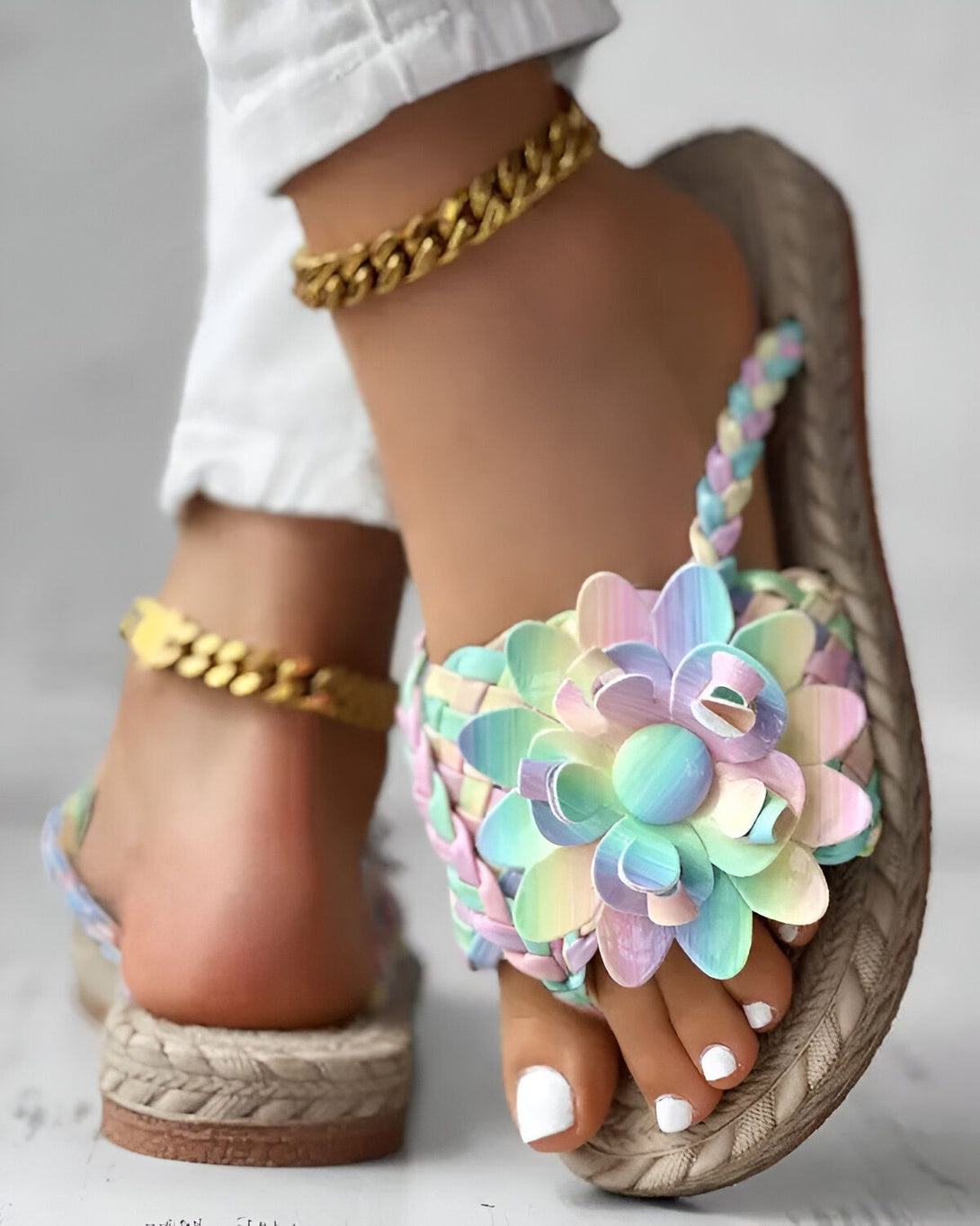 Une femme porte des sandales plates à fleurs colorées. Les sandales sont faites de lanières de cuir tressées et la semelle est en corde. La femme porte également une chaîne dorée autour de la cheville.