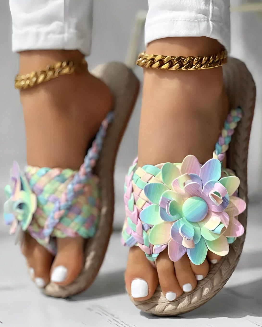Une femme porte des sandales plates à fleurs colorées. Les sandales sont faites de paille tressée et les fleurs sont en plastique. La femme a les ongles des pieds peints en blanc et porte un bracelet de cheville doré.