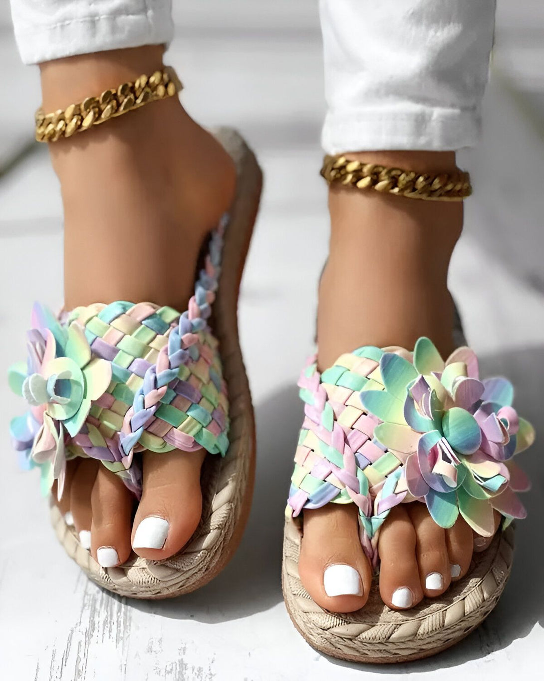 Une femme porte des tongs plates à fleurs multicolores. Les tongs sont faites de paille et ont une semelle compensée. La femme porte également un pantalon blanc et des bijoux dorés.
