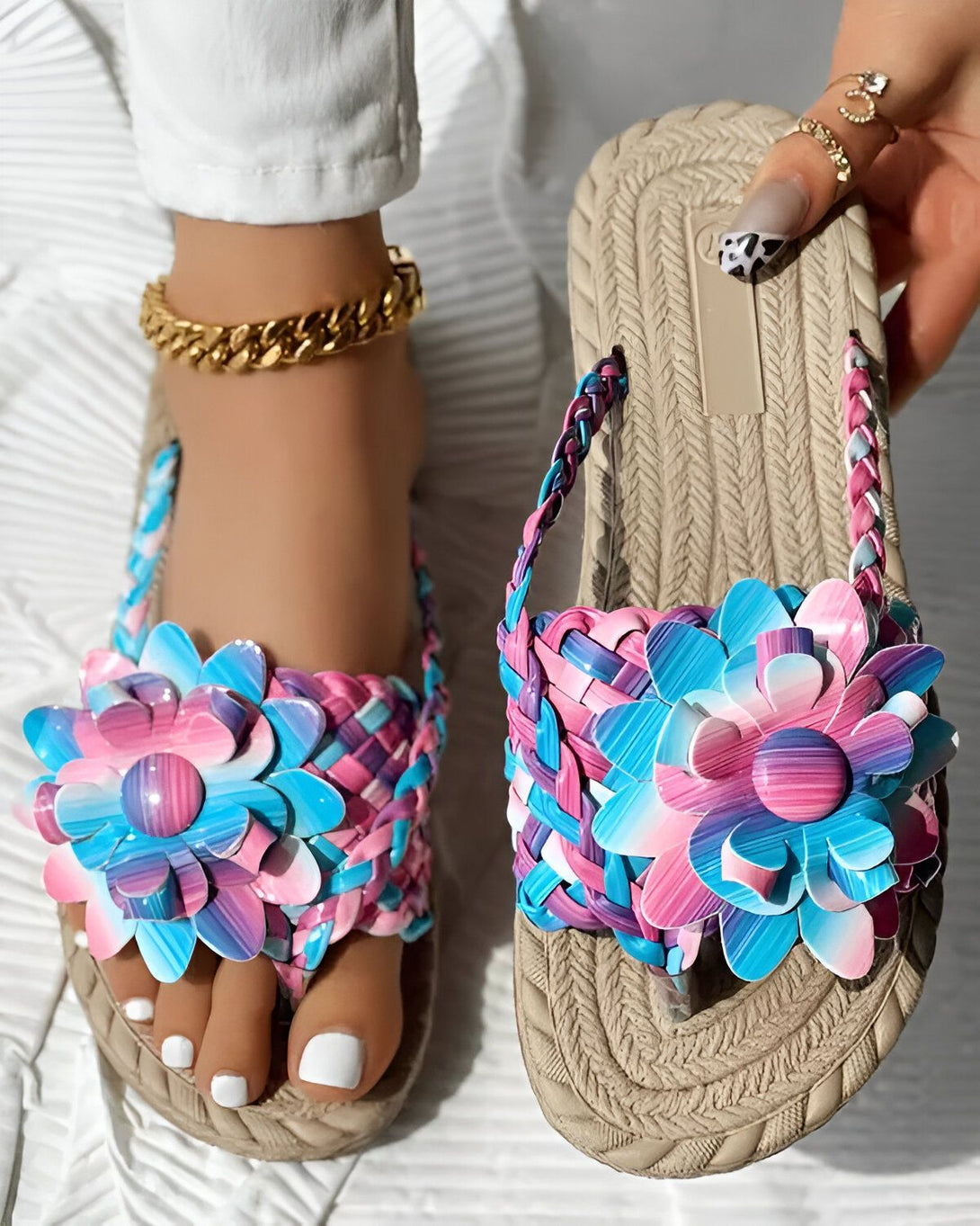Une femme porte des sandales plates avec des fleurs colorées. Les sandales sont beige et les fleurs sont roses, bleues et violettes. La femme a les ongles des pieds peints en blanc et porte un bracelet de cheville doré.