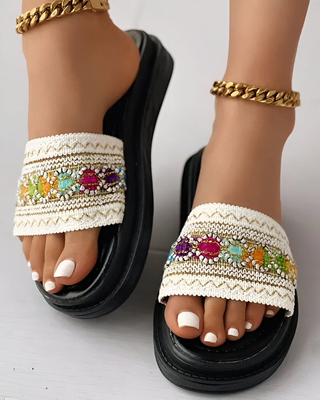 Une paire de sandales plates blanches avec des perles colorées. Les sandales sont portées par une femme qui a les ongles des pieds peints en blanc. La femme porte également un bracelet de cheville en or.