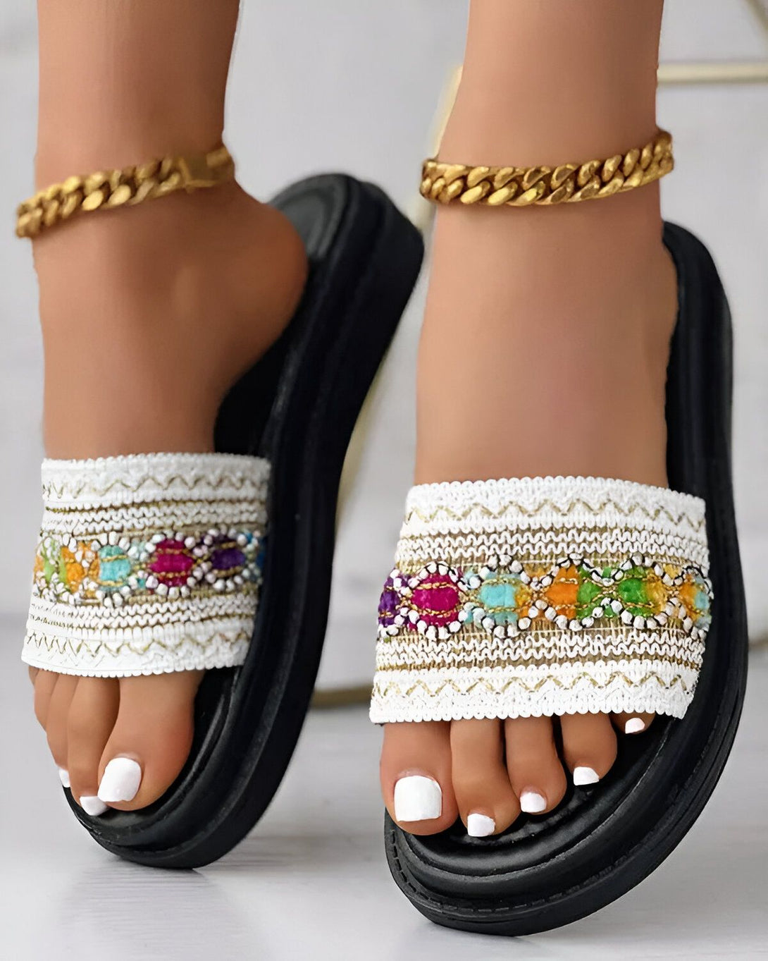 Une femme porte des sandales plates blanches avec des perles colorées. Les sandales ont une semelle noire et sont portées avec un pantalon blanc et un haut coloré.
