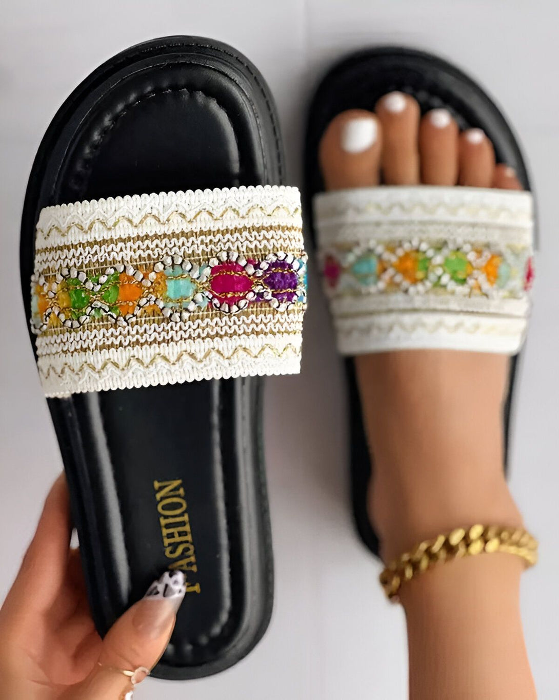 Une paire de sandales plates noires avec une bride blanche brodée de motifs colorés. Les sandales sont portées par une femme qui a les ongles des pieds peints en rose.