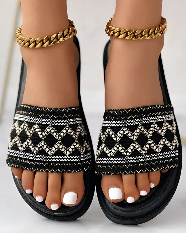 Une paire de sandales plates noires et blanches avec un motif de losanges. Les sandales sont portées par une femme qui a les ongles des pieds peints en blanc et qui porte un bracelet de cheville en or.