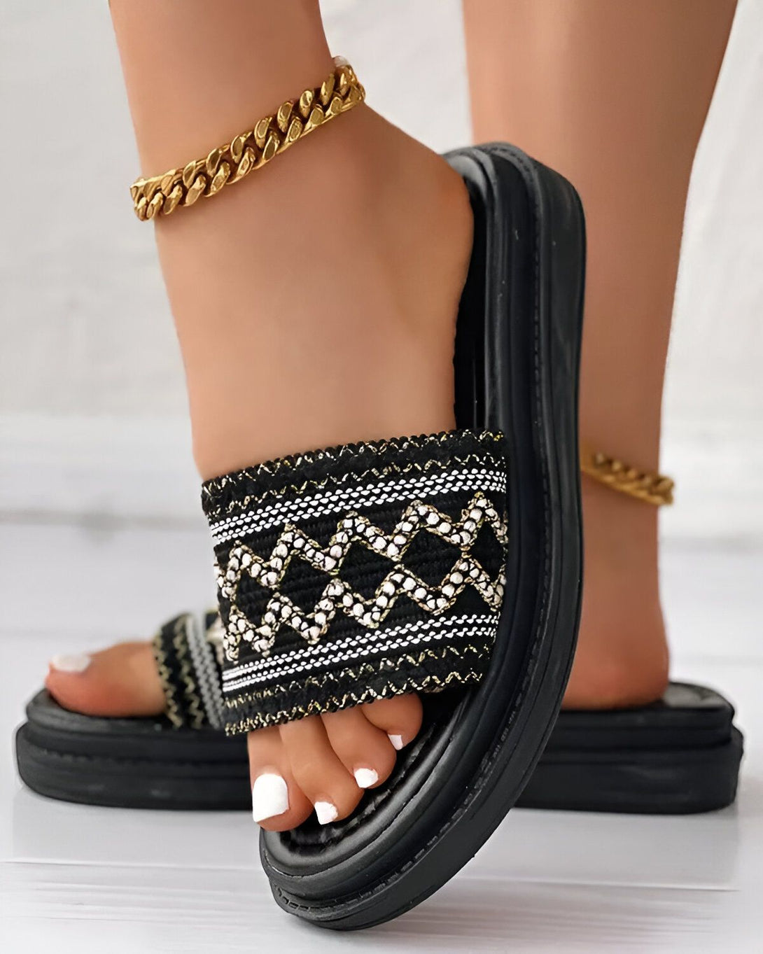 Une femme porte des sandales noires et blanches à motifs géométriques. Les sandales ont une semelle épaisse et sont ornées d'une chaîne dorée. La femme porte également un bracelet de cheville doré.