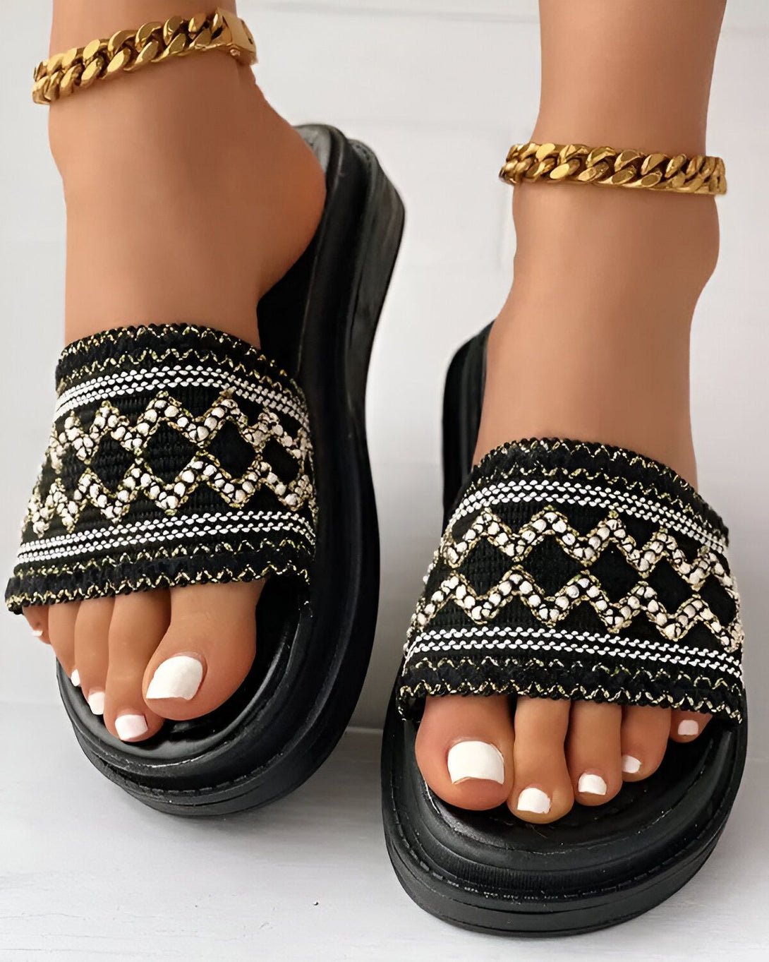 Une paire de sandales noires à enfiler avec un motif ethnique coloré sur le dessus. Les sandales sont portées par une femme qui a les ongles des pieds peints en blanc.