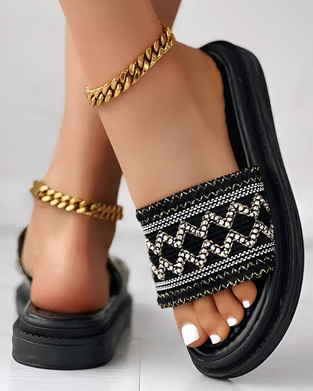 Une femme porte des sandales noires et dorées. Les sandales sont plates et ont une bride large. La bride est décorée d'un motif géométrique noir et doré. La semelle est noire et épaisse. La femme a les ongles des pieds peints en blanc. Elle porte également un bracelet de cheville doré.