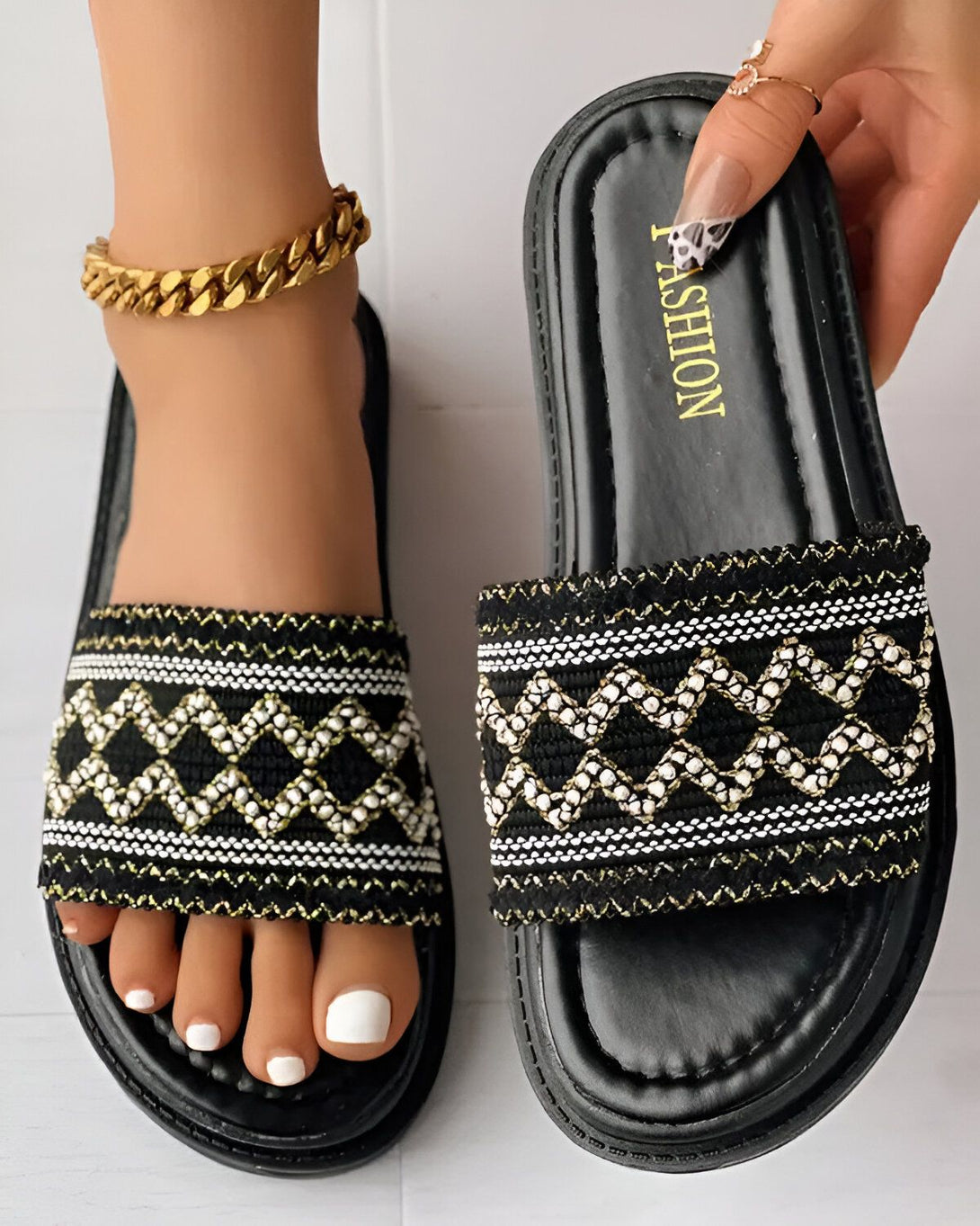 Une femme porte des sandales noires et blanches. Les sandales sont plates et ont une bride large. La femme a les ongles des pieds peints en blanc. Elle porte également un bracelet de cheville en or.