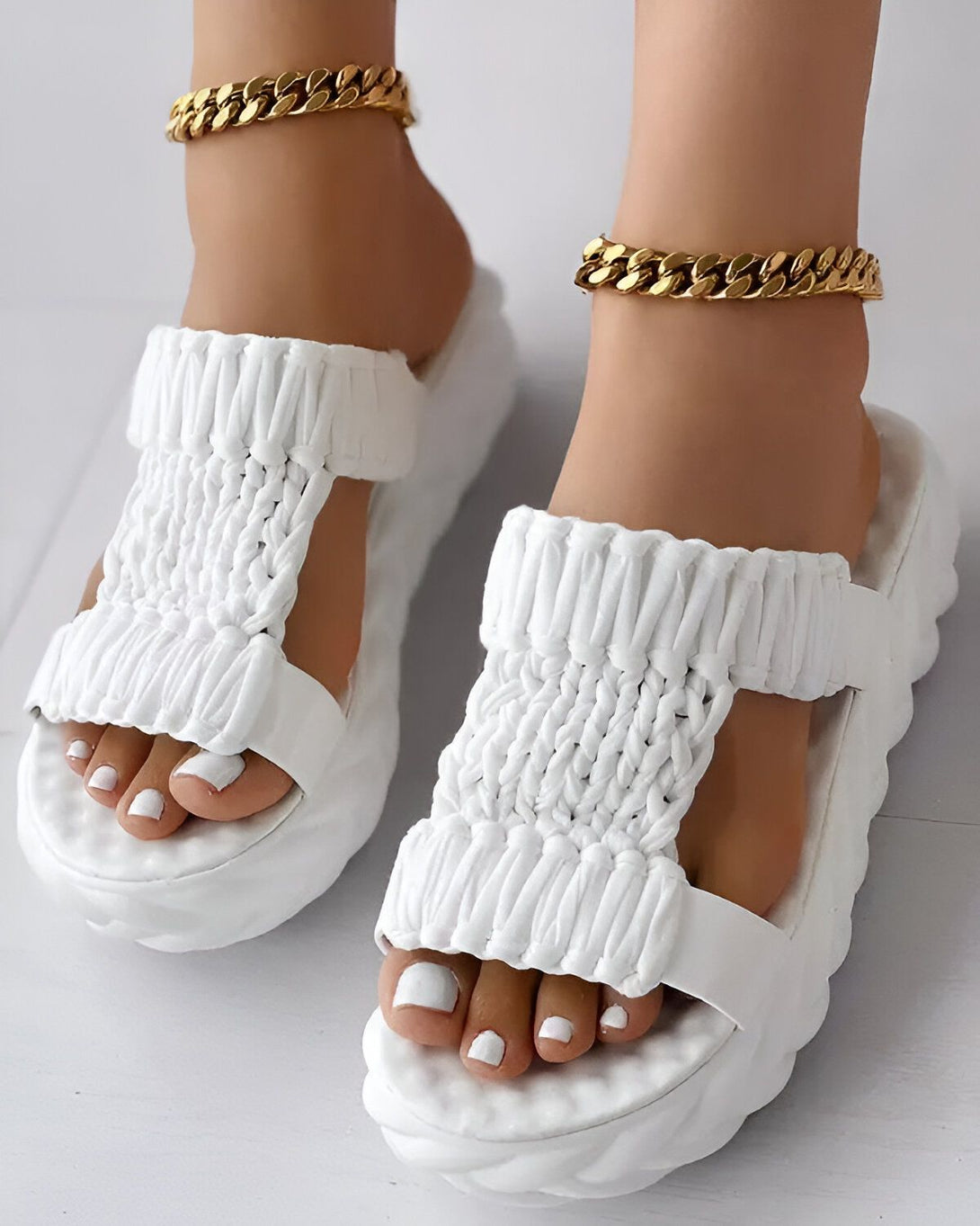 Une paire de sandales blanches à plateforme pour femme. Les sandales sont dotées de deux brides croisées sur le dessus du pied et d'une semelle épaisse. La femme porte également une chaîne dorée à la cheville.