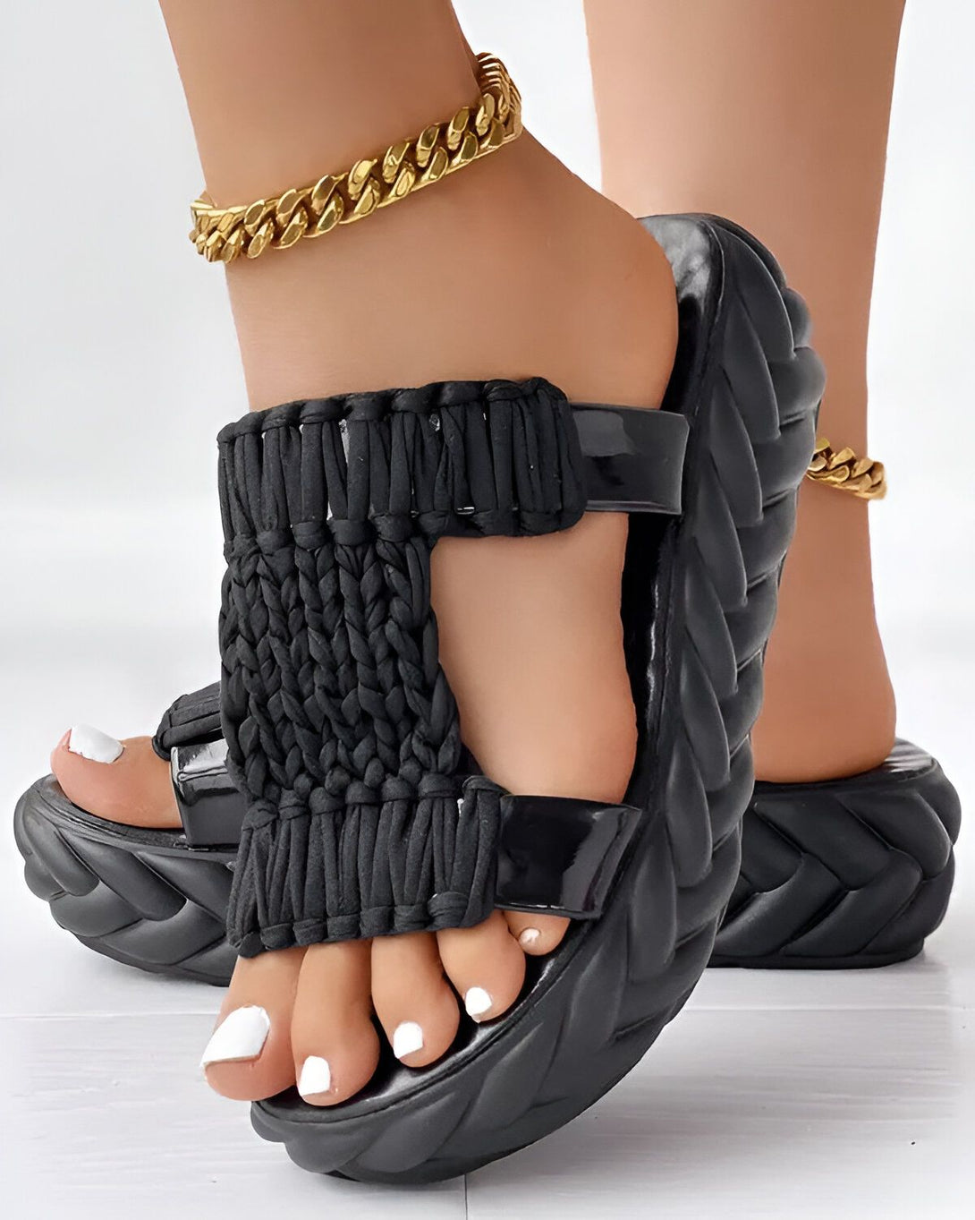 Une femme porte des sandales noires à talons compensés. Les sandales sont en matière synthétique et ont une bride tressée sur le dessus. La femme a les ongles des pieds peints en blanc et porte un bracelet de cheville en or.