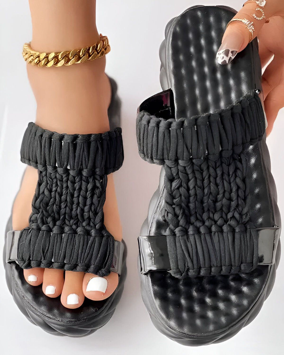 Une femme porte des sandales noires avec des lanières tressées. Les sandales ont une semelle épaisse et une bride qui passe entre les orteils. La femme a les ongles des pieds peints en blanc et porte un bracelet de cheville en or.