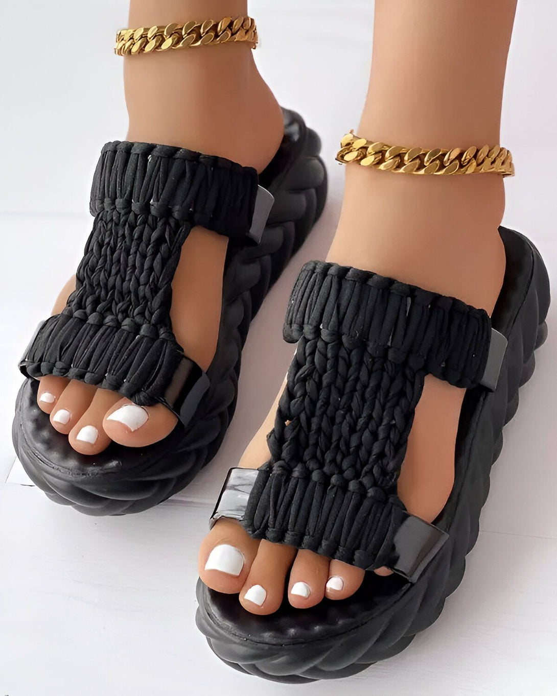 Une femme porte des sandales noires avec des semelles épaisses. Les sandales sont faites d'un matériau tressé et ont des brides velcro. La femme a les ongles des pieds peints en blanc et porte des bracelets de cheville en or.