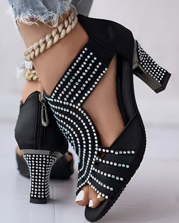 Une image d'une paire de sandales à talons hauts noires avec des strass. Les sandales sont portées par une femme qui porte un jean bleu et un haut noir. La femme est debout sur un sol en marbre blanc.