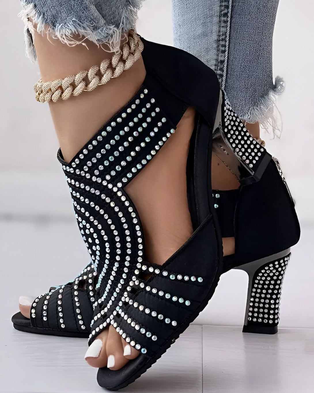 Une femme porte des sandales noires à talons hauts. Les sandales sont recouvertes de strass et ont une bride à la cheville. La femme porte un jean bleu et un bracelet doré à la cheville.