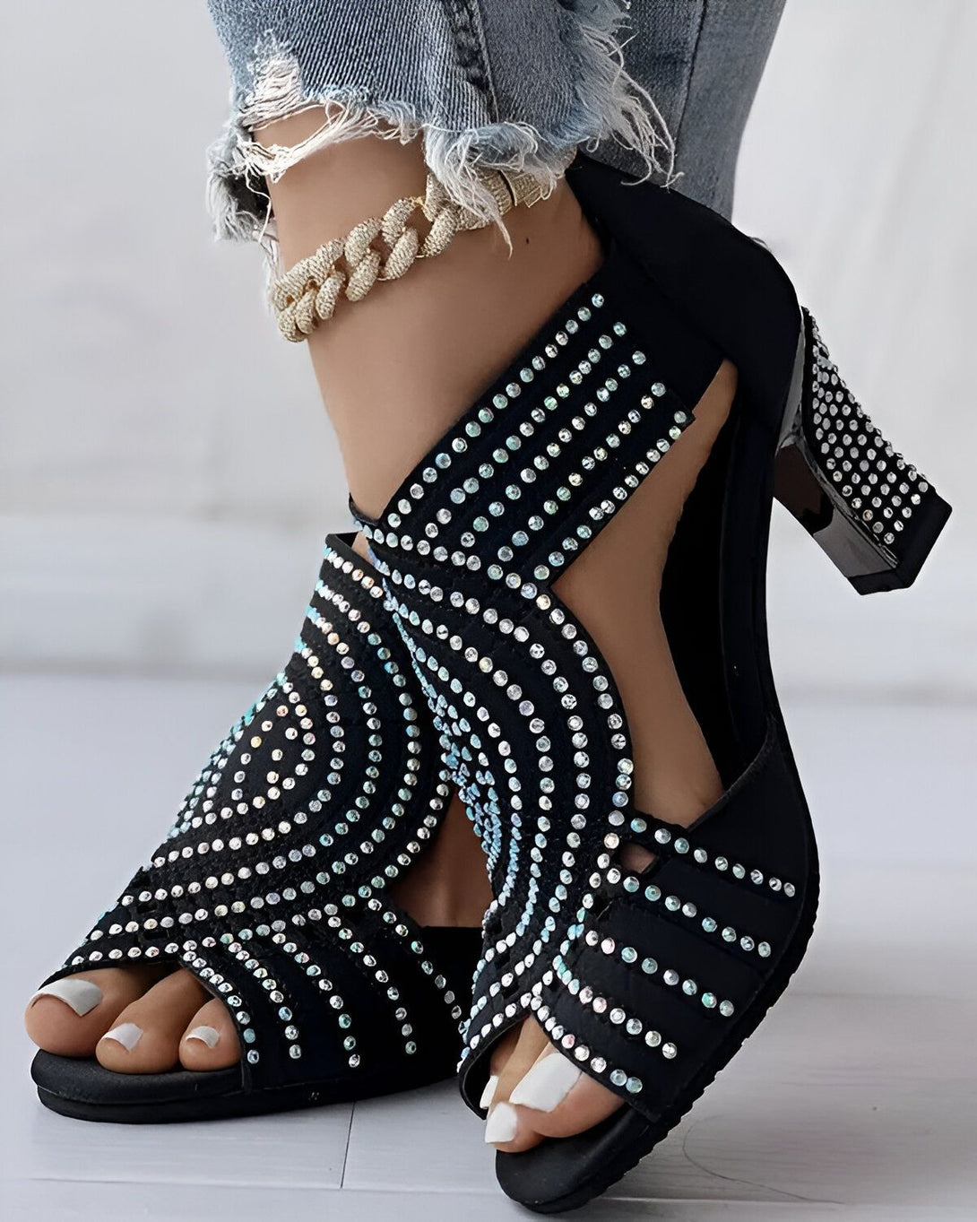 Une femme porte des sandales noires à talons hauts. Les sandales sont recouvertes de strass argentés. La femme porte un jean bleu déchiré et un chemisier blanc. Elle est debout sur un sol en marbre blanc.