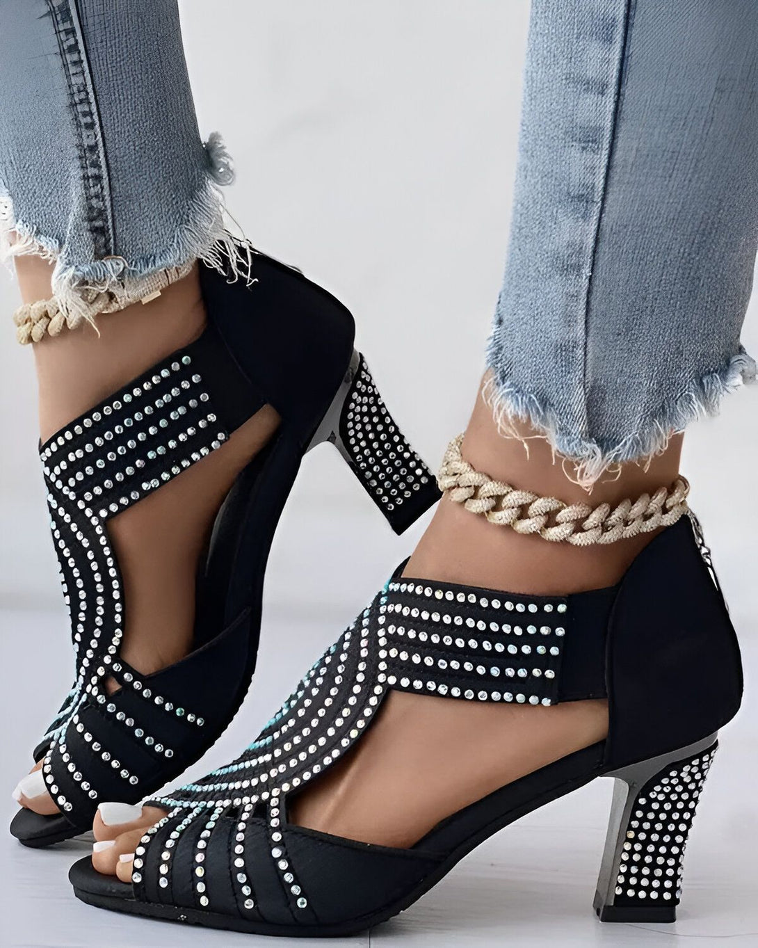 Une femme porte des sandales noires à talons hauts. Les sandales sont ornées de strass. La femme porte un jean bleu déchiré.