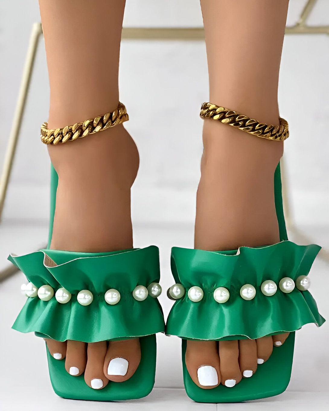 Une femme porte des sandales vertes à talons hauts avec des perles et des volants. Les sandales sont en cuir et ont une semelle verte. La femme a les ongles des pieds peints en blanc et porte des bracelets de cheville en or.