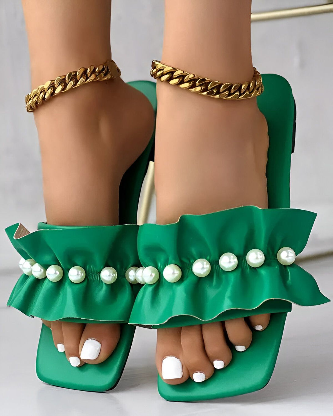 Une femme porte des sandales vertes à talons hauts avec des perles et des volants. Les sandales sont en cuir et ont une semelle verte. La femme a les ongles des pieds blancs et porte des bracelets de cheville en or.