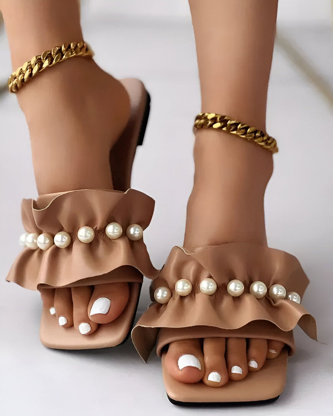 Une paire de sandales marron clair à talons hauts avec des perles et des volants sur le dessus. Les sandales sont portées par une femme avec une peau bronzée et des ongles blancs. La femme porte également une chaîne en or autour de la cheville.