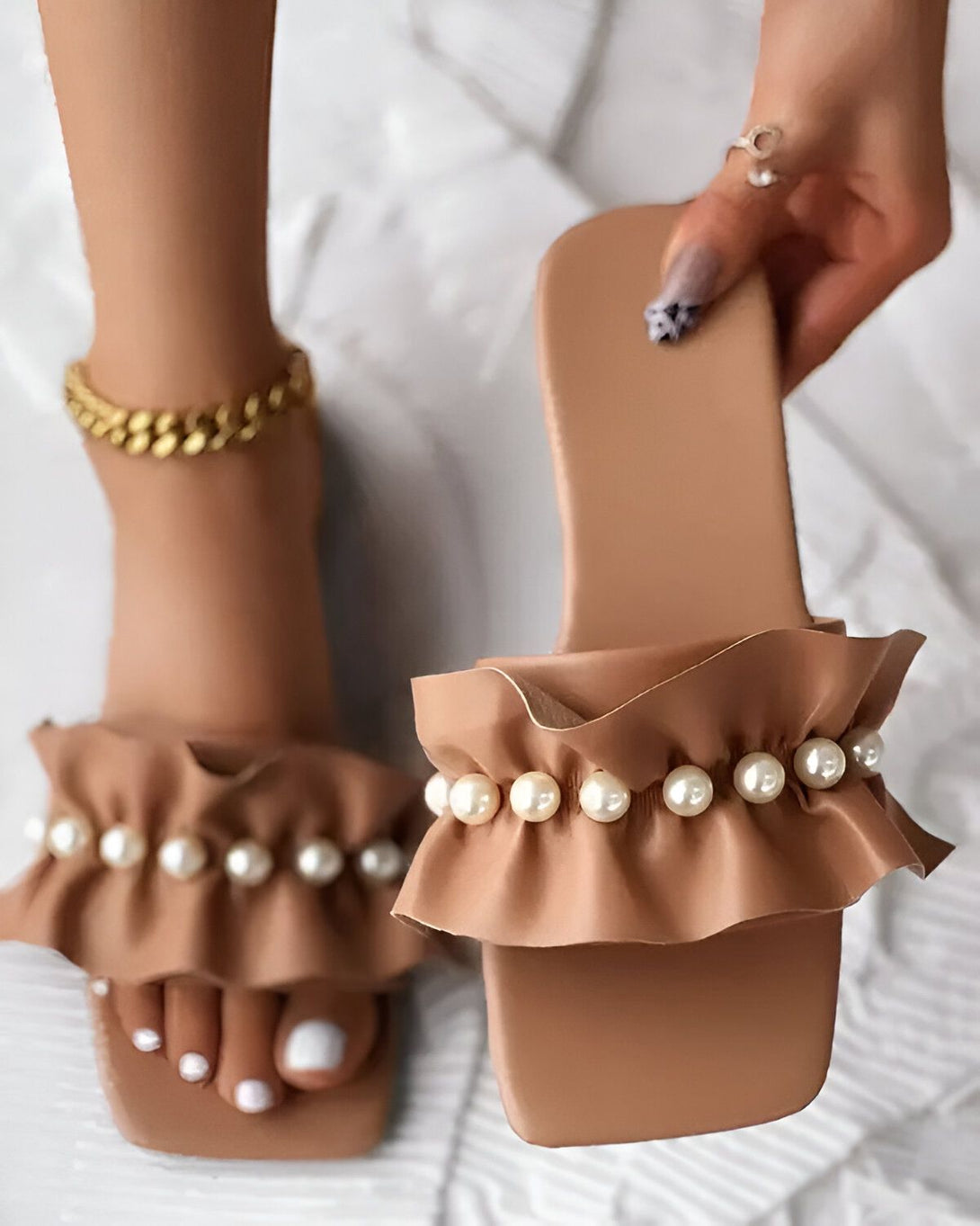 Une femme porte des sandales marron clair à talons carrés. Les sandales sont ornées de perles et de volants. La femme a les ongles des pieds peints en blanc. Elle porte également un bracelet de cheville en or.