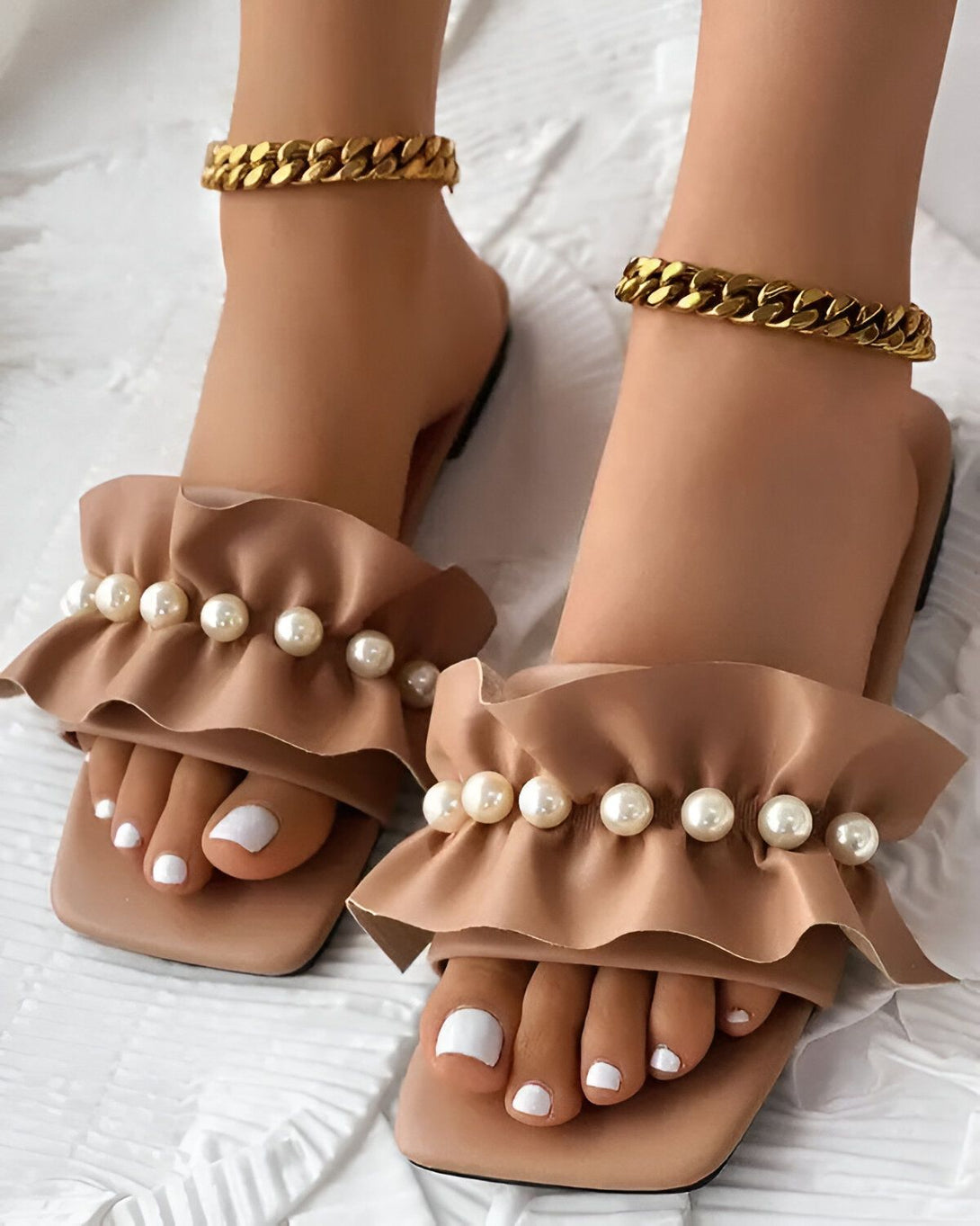 Une paire de sandales marron clair à talons carrés avec des perles et des volants sur le dessus. Les sandales sont portées par une femme qui a les ongles des pieds peints en blanc et qui porte des bracelets de cheville en or.