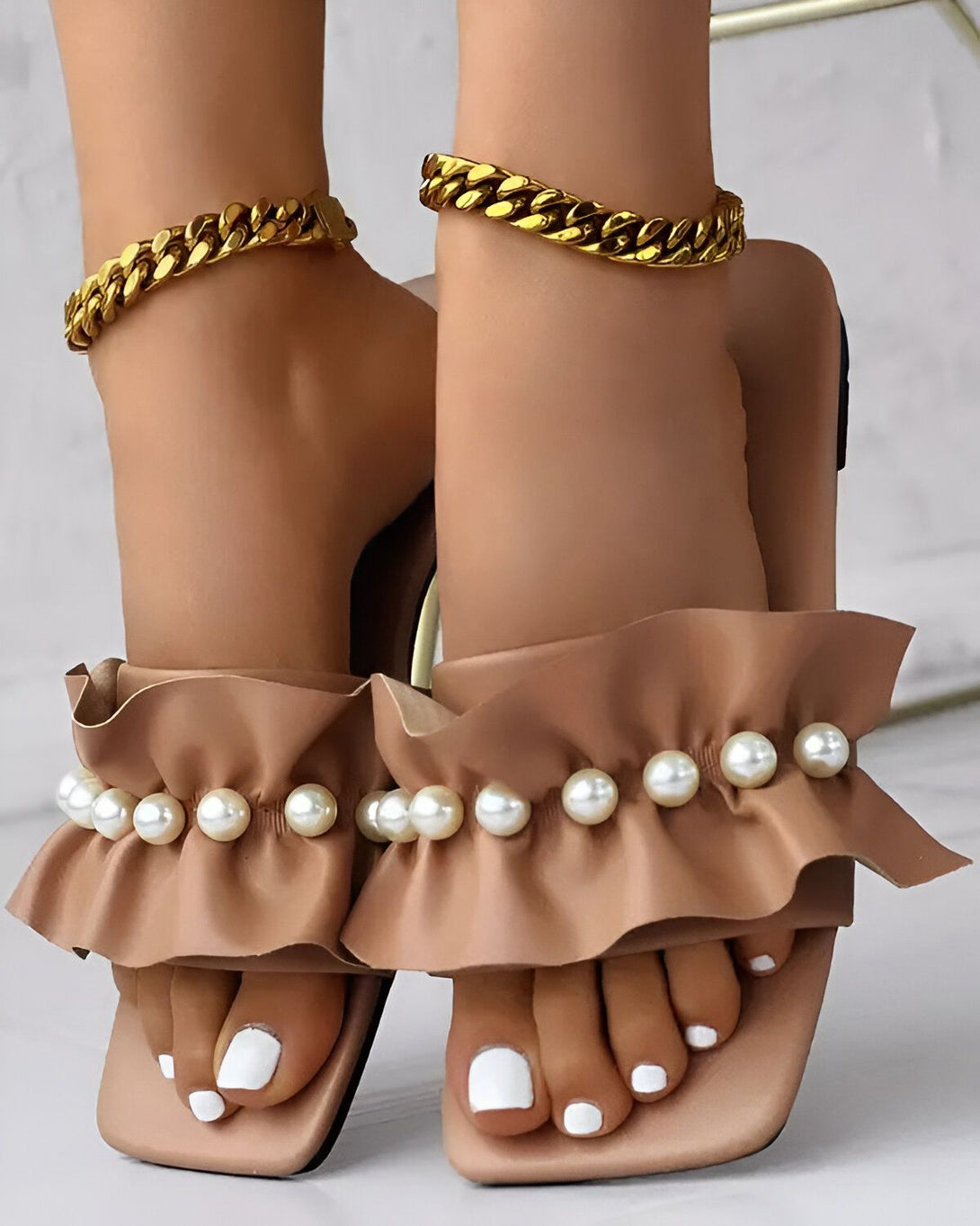Une paire de sandales marron clair à talons hauts avec des perles et des volants sur le devant. Les sandales sont portées par une femme qui a les ongles des pieds peints en blanc et qui porte des bracelets de cheville en or.