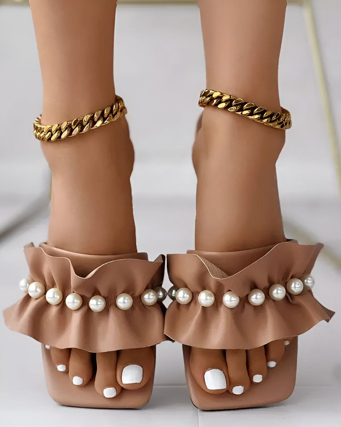Une paire de sandales à talons hauts de couleur beige avec des perles et des volants. Les sandales sont portées par une femme qui a les ongles des pieds peints en blanc et qui porte des bracelets de cheville en or.