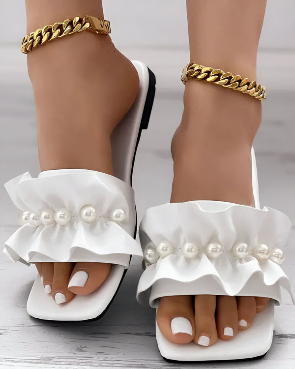 Une paire de sandales blanches à talons avec des perles et des volants. Les sandales sont portées par une femme qui a les ongles des pieds peints en blanc. Elle porte également un bracelet de cheville en or.