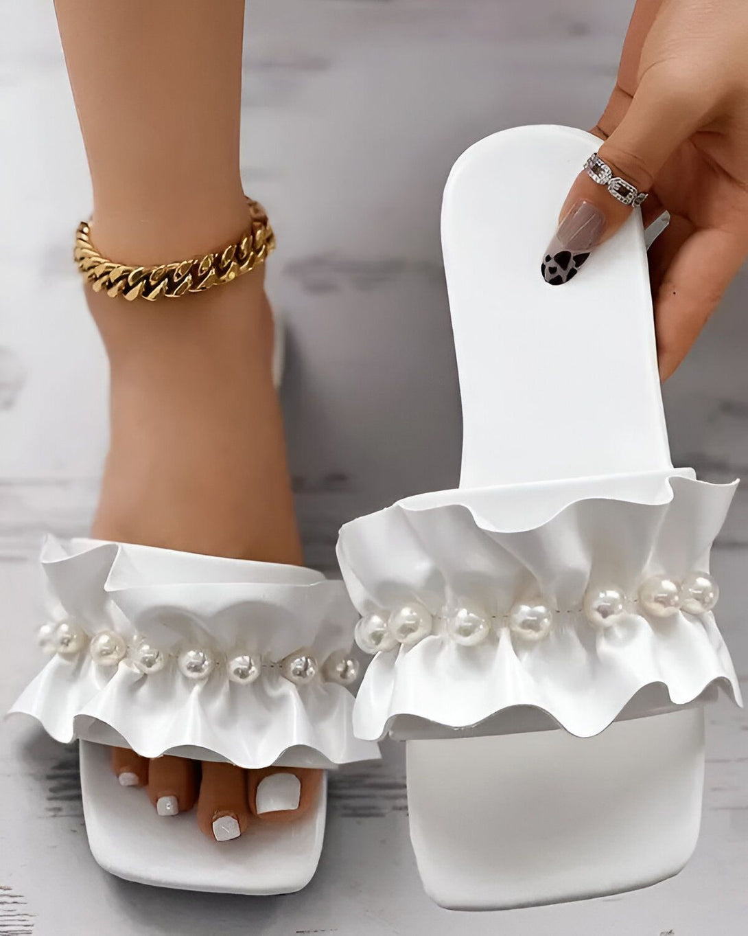 Une femme porte des sandales blanches à talons hauts avec des perles et des volants. Les sandales sont ouvertes au niveau des orteils et ont une bride qui s'enroule autour de la cheville. La femme a les ongles des pieds peints en blanc et porte un bracelet de cheville en or.