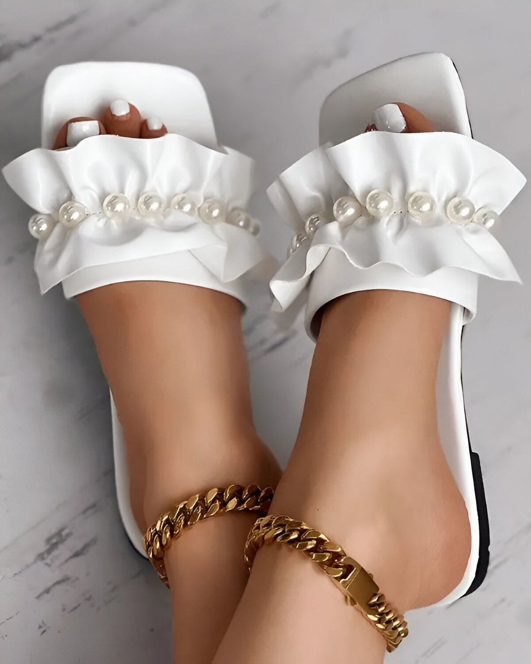 Une paire de sandales blanches à talons hauts avec des perles et des volants. Les sandales sont portées par une femme qui a les ongles des pieds peints en blanc. Elle porte également un bracelet de cheville en or.