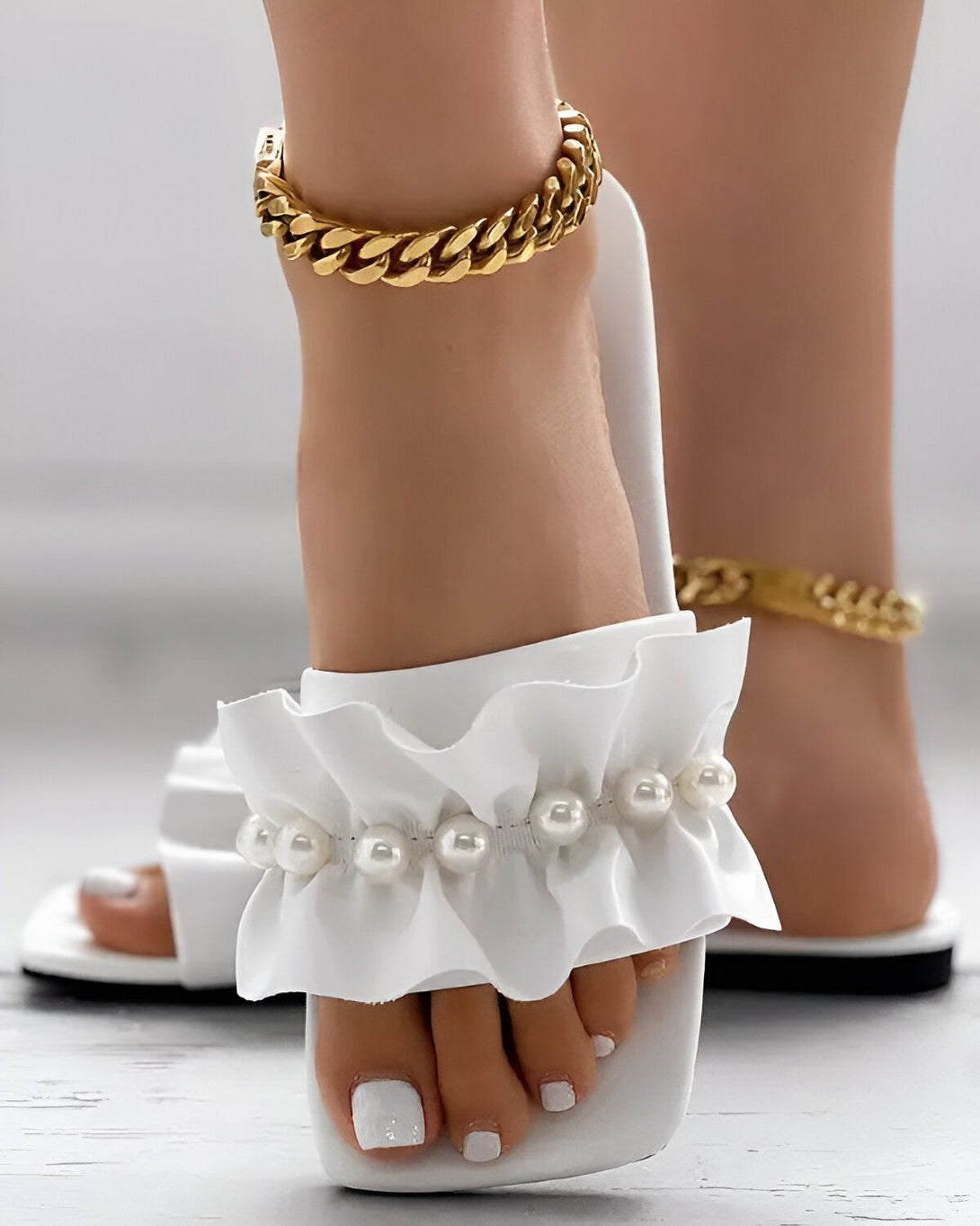 Une femme porte des sandales blanches à talons hauts avec des perles et des volants. Elle porte également une chaîne dorée à la cheville.