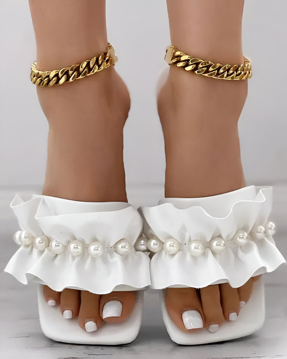 Une paire de sandales blanches à talons hauts avec des perles et des volants. Les sandales sont portées par une femme qui a les pieds nus et qui porte des bracelets de cheville en or.