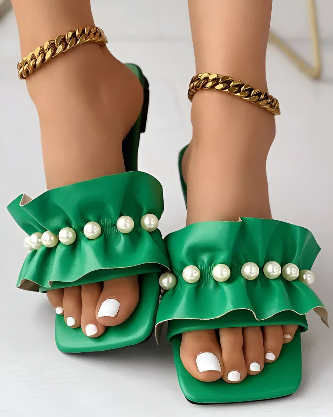 Une paire de sandales plates vertes à bout carré avec des perles blanches sur le dessus. Les sandales sont portées par une femme qui a les ongles des pieds blancs et qui porte des chaînes dorées aux chevilles.