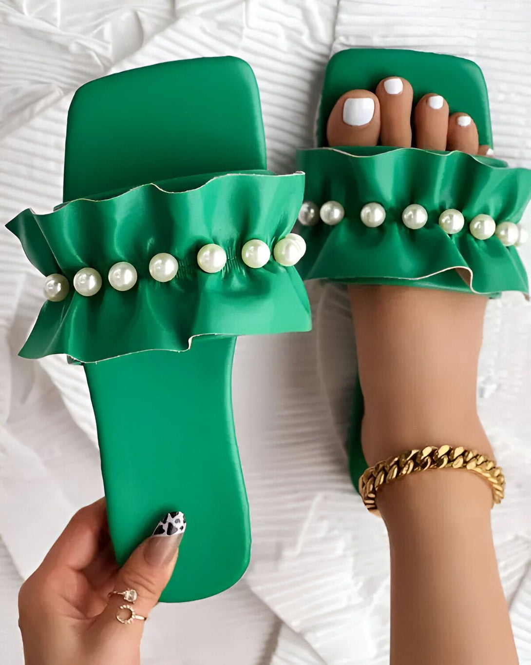 Une femme porte des sandales vertes à talons hauts. Les sandales sont ornées de perles blanches. La femme a les ongles des pieds peints en blanc. Elle porte également un bracelet de cheville en or.