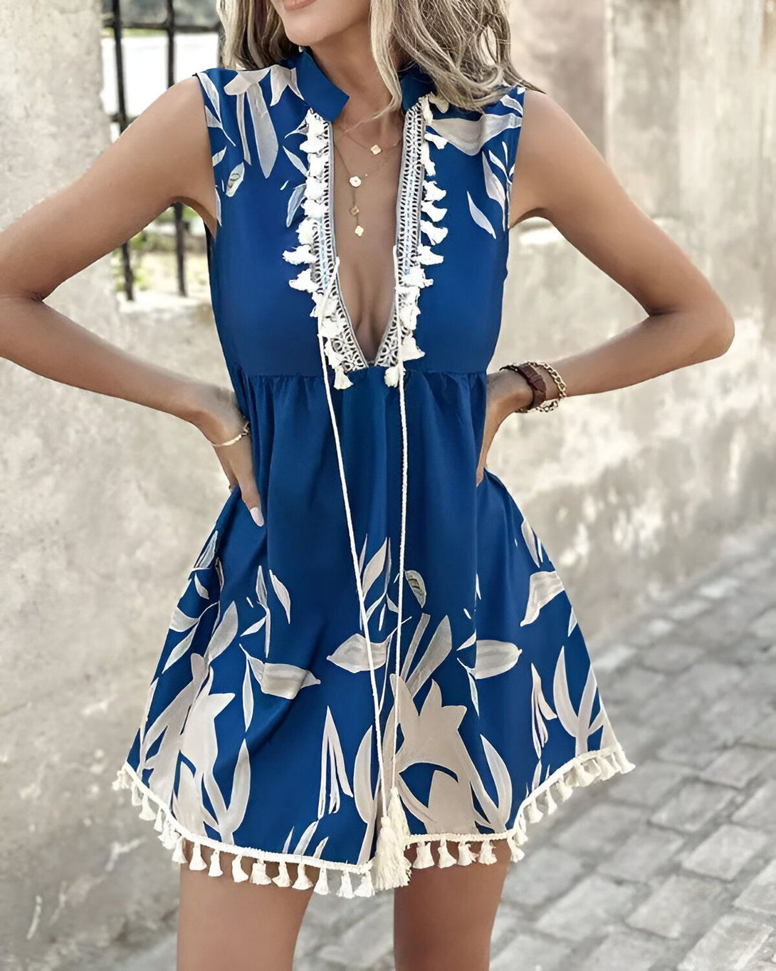 Une femme porte une robe bleu marine sans manches à imprimé de feuilles tropicales. La robe est courte et évasée, avec un col montant et des pompons blancs sur la poitrine. La femme est blonde et porte un collier doré. Elle est debout devant un mur de pierre.