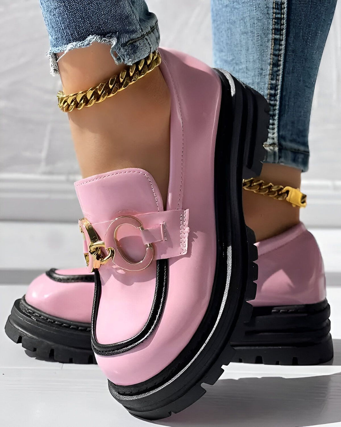 Une femme porte des mocassins roses avec des semelles épaisses noires. Les mocassins ont des boucles dorées. La femme porte également un jean bleu et des chaussettes blanches.